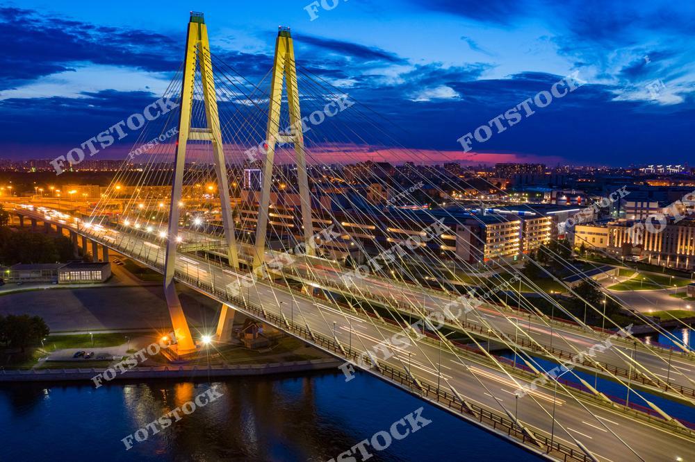 На фотографии изображен Большой Обуховский мост, который пересекает реку Неву и является одним из самых известных вантовых мостов Санкт-Петербурга. Ночное освещение подчеркивает архитектурную конструкцию моста, а огни города отражаются в водах реки. Этот мост — один из немногих, который не разводится, обеспечивая круглосуточное автомобильное движение между берегами. Вид с высоты открывает панораму ночного Санкт-Петербурга.